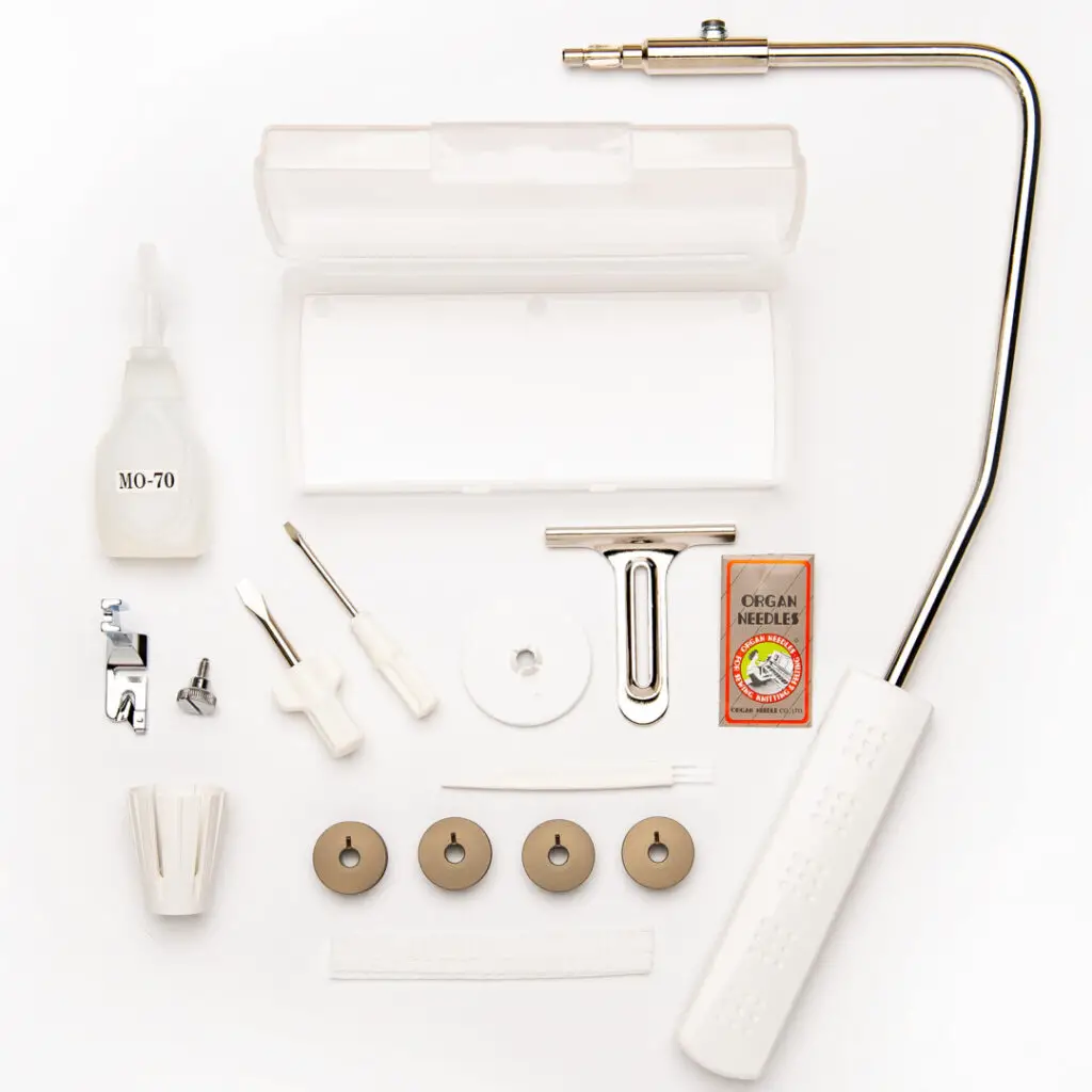 The additional accessories laid out, knee lifter, four bobbins, hemmer foot, screwdriver, oil, cloth guide, spool cap, lint brush, spool net, needles, and accessory box.