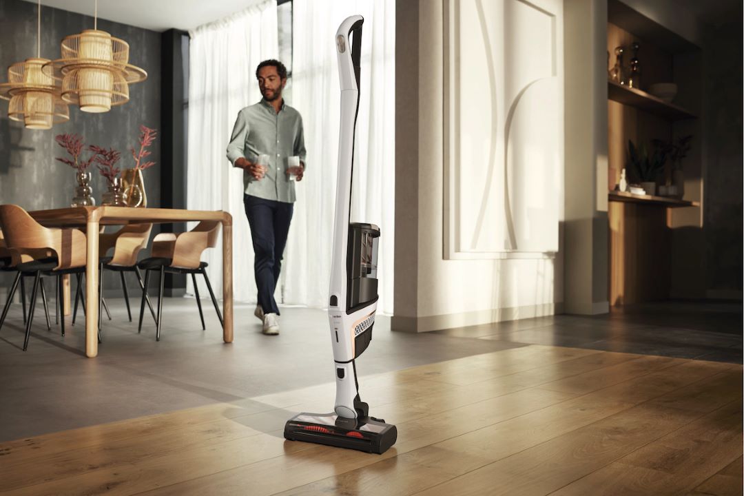 H2X cordless vacuum standing upright in a room with hardwood floors and a man in the background.