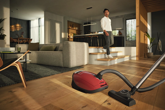 In the living room of a modern home a Miele vacuum rests on hardwood floors ready to use as a man walks towards it.