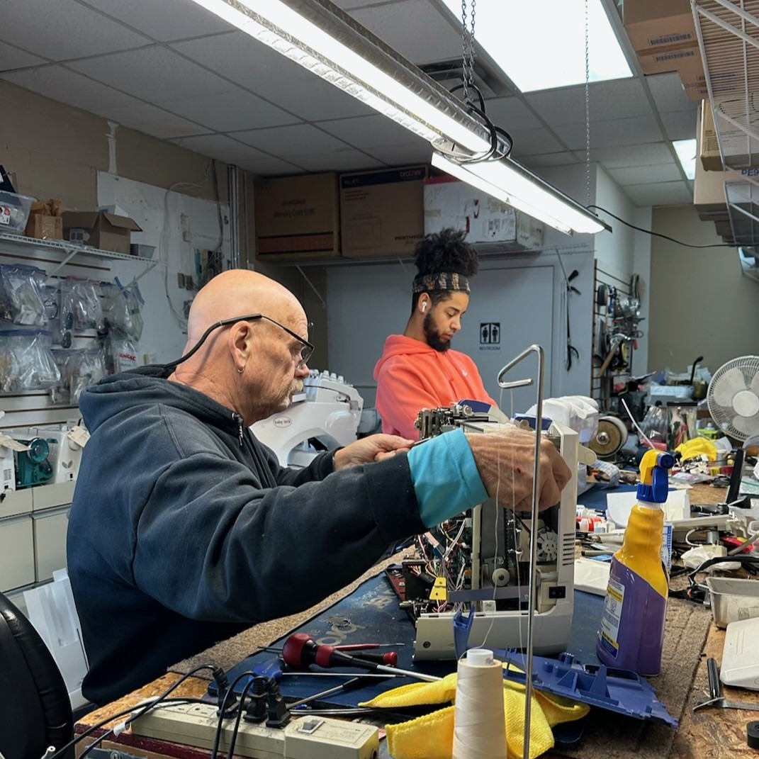 Two sewing machine repair technicians sit at a workbench inside a repair shop. Tools including threads, scissors, and wrenches are spread across the desk. In the foreground, Johnny is actively disassembling a sewing machine.